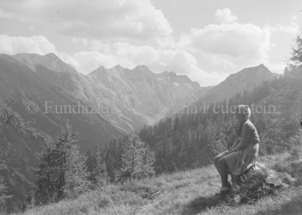 Frau auf Baumstumpf sitzend, Wald und Berge dahinter