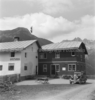 Gasthaus, Automobil und Tische davor, Berge hinten