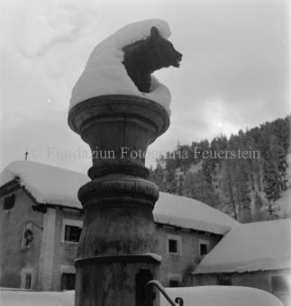 Schneebedeckte Bärenskulptur auf Brunnen, dahinter Häuser