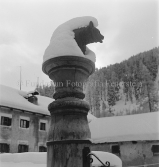 Schneebedeckte Bärenskulptur auf Brunnen, dahinter Häuser