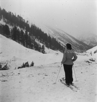Skitourengängerin, auf Berghang blickend, hinten Dorf