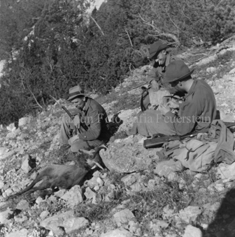 Jäger an Berghang sitzend, geschossene Gämse mit Zweig im Mund 