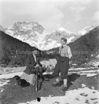 Mann auf Stein sitzend, mit Gewehr und Hund, Frau daneben stehend, Bergpanorama, teilweise Schnee