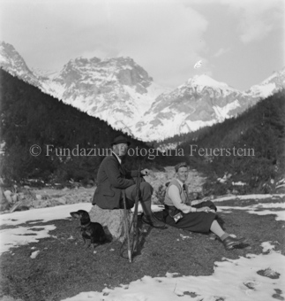 Frau auf Boden, Mann auf Stein sitzend, mit Gewehr und Hund, Bergpanorama, teilweise Schnee