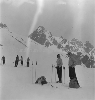 Gruppe Skitourengänger im Schnee stehend, Skistöcke in Boden, hinten Berggipfel