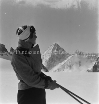 Skitourengänger von der Seitem Skistöcke in der Hand, Brille auf Stirn, hinten Berggipfel
