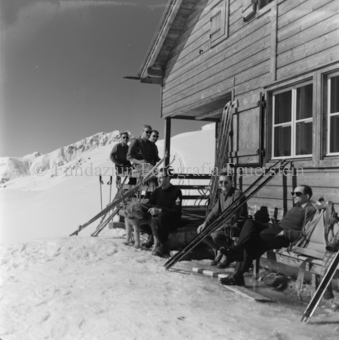 Skitourengängergruppe vor Berhütte sitzend, Ski an Wand, viele mit Sonnenbrille