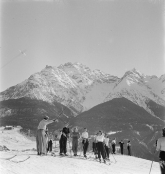 Gruppe Skifahrerinnen, Mann Skistock in Richtung weisend, hinten Berg 
