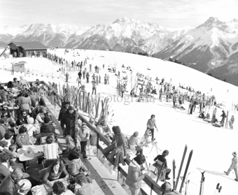 Restaurantterrasse auf Berghütte, viele Skifahrer und Personen zu Tisch, schneebedeckte Berglandschaft im Hintergrund