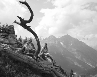 Zwei Frauen auf altem Arvenstamm neben Steinmauer, Berglandschaft