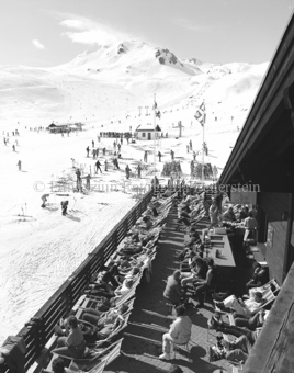 Personen auf Sonnenterrasse vor «Fredl's Eisbar und Grillstation», Skifahrer bei Talstation, Schlepplift, Fahnen