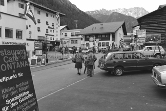 Personen und Autos auf Parkplatz und Strasse im Dorf, Restaurants, Kantonalbank, und Läden