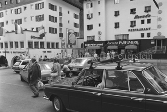 Personen und Autos auf Parkplatz und Strasse im Dorf, Restaurants, Kantonalbank, und Läden