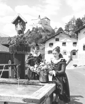 Zwei Frauen in Tracht mit Blumen beim Brunnen, Schloss Tarasp im Hintergrund