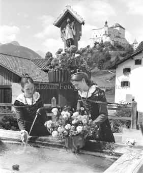 Zwei Frauen in Tracht mit Blumen beim Brunnen, Schloss Tarasp im Hintergrund