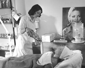 Kosmetikerin benutzt Dampfgerät an Frau unter Tüchern auf Liege, Kosmetika im Hintergrund