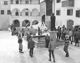L'Hom Strom, Personen bauen Strohmann auf Dorfplatz neben Brunnen, Fassade mit Sgraffito