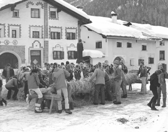L'Hom Strom, Personen bauen Strohmann auf Dorfplatz neben Brunnen, Fassade mit Sgraffito