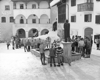 L'Hom Strom, Personen bauen Strohmann auf Dorfplatz neben Brunnen, Fassade mit Sgraffito