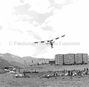 Hängegleiterflieger über dem Boden im Tal, Zuschauer mit Autos, Dorf im Hintergrund