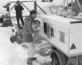 Mann lädt Husky Schlittenhund in Transportkiste auf Anhänger mit Schlitten, zwei Kinder und Fotograf im Hintergrund