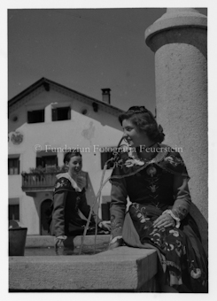 Zwei Frauen in Tracht an brunnen, Wohnhaus im Hintergrund