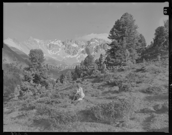 Porträt Frau auf Bergwiese neben Alpenrosen sitzend, Nadelbäume und Berggipfel im Hintergrund