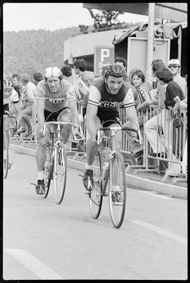 Drei Velo-Rennfahrer in Fahrt, auf dem Trottoir neben einem Parkhaus Zuschauer 