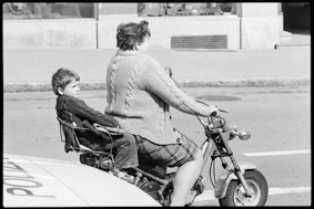 Frau auf Motorrad mit Kind auf dem Rücksitz, daneben ein Polizeiauto