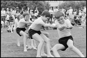 Kinder in Turnkleidung am Seilziehen auf Rasen, im Hintergrund Zuschauende