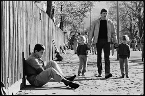 Ein Waffenläufer sitzt am Boden, daneben ein Passant mit zwei Kindern, im Hintergrund Sportler und Waffenrucksäcke