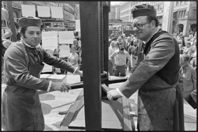 zwei Männer in Kleidung von früher bei der Arbeit mit einer Papierpresse, zum Trocknen aufgehängte Papiere, im Hintergrund Schaulustige