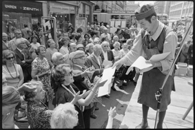 Mann in Kleidung von früher beim Verteilen von geschöpftem Papier an Schaulustige, im Hintergrund Tram bei der Station Bärenplatz