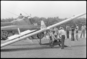 einplätziges Flugzeug F-AZBJ am Boden stehend, ein Mann setzt sich auf den Sitz, daneben Schaulustige und ein Panzer