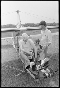 einplätziges Flugzeug F-AZBJ am Boden stehend, ein Mann sitzt darauf, ein Kind und ein Mann schauen zu