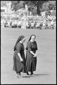 Zuschauer auf  einer grossen Wiese um einen Platz auf Bänken sitzend, vorne zwei Klosterfrauen, im Hintergrund ein Glace-Stand