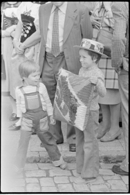 Kind mit Berner-Wappen-Kissen, auf dem Kopf Hut mit Berner Wappen und Béliers-Logo, daneben Besucher des Anlasses und Trachtenfrauen