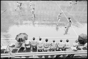 Matchszene auf dem «Fussball-Feld», daneben die Ersatzbank mit Fussballspielern und Regenschirmen