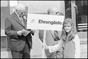 Mädchen mit einem Hinweis-Schild, darauf das Logo vom Eidg. Musikfest Biel 1976 und die Aufschrift «Ehrengäste», daneben zwei Gäste mit Festabzeichen