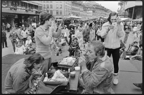 Schauspieler auf einer Bühne sitzend beim öffentlichen Schminken, daneben Zuschauende auf Stühlen, im Hintergrund Marktstände