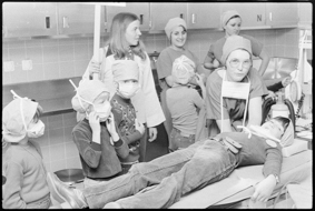 Ärztin, Lehrerin und Kinder in OP-Kleidung, eines der Kinder liegt auf dem Operationsstisch