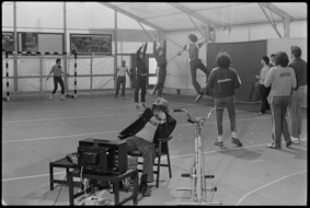 Schauspieler vor Fernseher sitzend, dahinter Handballspieler