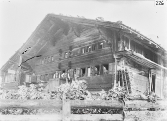 Zweistöckiges Holzgebäude mit geschindeltem Satteldach giebelseitigem, zentalem, Eingang, zwei Männer an Brüstung stehend, rechts von ihnen 3 Frauen und ein Kind aus den Fenstern schauend, Blockkonsolen und Fries, Im Vordergrund Bretterzaun