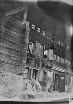 Dreistöckiges Wohngebäude mit gemauertem Erdgeschoss, von Blockkonsolen getragenes Dach, Fassade mit Friesen und Holzschnitzereien, ältere Frau aus Fenster im 1. OG schauend, Frau und Mädchen vor Fenster in Erdgeschoss sitzend