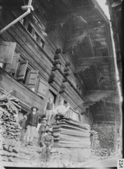 Giebelseite dreistöckiges Holzgebäude mit von Blockkonsolen getragenem Satteldach vor dem Gebäude aufgestapelte Holzbohlen und Brennholz, sechs Knaben 