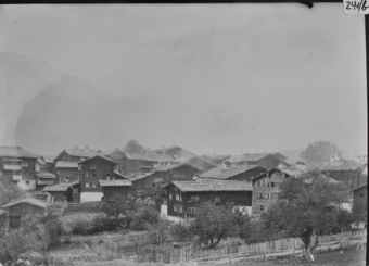 Dorf in flachem Bergthal Im Vordergrund  Obstbäume, Bretter- und Lattenzäune, mehrstöckige Holzgebäude mit gemauertem Erdgeschoss