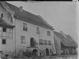 Dreistöckiges, gemauertes Wohngebäude mit geziegeltem Pultdach und Zinnengiebel, Eingang im 1. OG durch gemauerte Treppe mit Korbbogen erschlossen, links Anbau mit Obergeschoss in Fachwerkbaauweise, rechts im Hintergrund Steingebäude Pultdach und Zinnengiebel, unter Dachvorspruch grosse Holzfässer