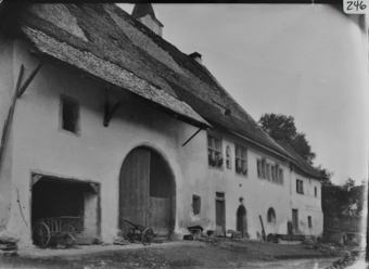 Traufseite zweistöckiges, gemauertes Bauernhaus mit steilem, geschindeltem Satteldach welches über Ökonomieteil durch Büge gestützt wird, darunter teils geöffnetes Tenntor, einspanniger Karren, deneben Sprossenwagen in Unterstand, Wohnteil mit rechteckigen Sprossenfester im OG, davor Brunnen und Hühner