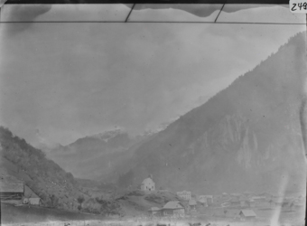 Bergthal mit kleiner Ortschaft und Kirche auf Hügel, im Hintergrund schneebedeckte Berggipfel