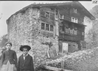 Dreistöckiges Bauernhaus an Hanglage mit massiver, thalseitiger Steinmauer und gezimmertem Wohnteil, geschindeltes Schleppdach, Klebdach über 1. OG, Butzen- und Sprossenfenster, kniehohe Steinmauer, links im Vordergrund ein Mädchen und ein Junge mit Hut, in die Kamera schauend